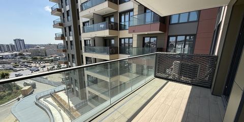 Yeni teslim edilmiş bir dairenin güneşli balkonu; cam korkuluk ve metal panelin ardında bloğun cephesi ve çevre mahalle